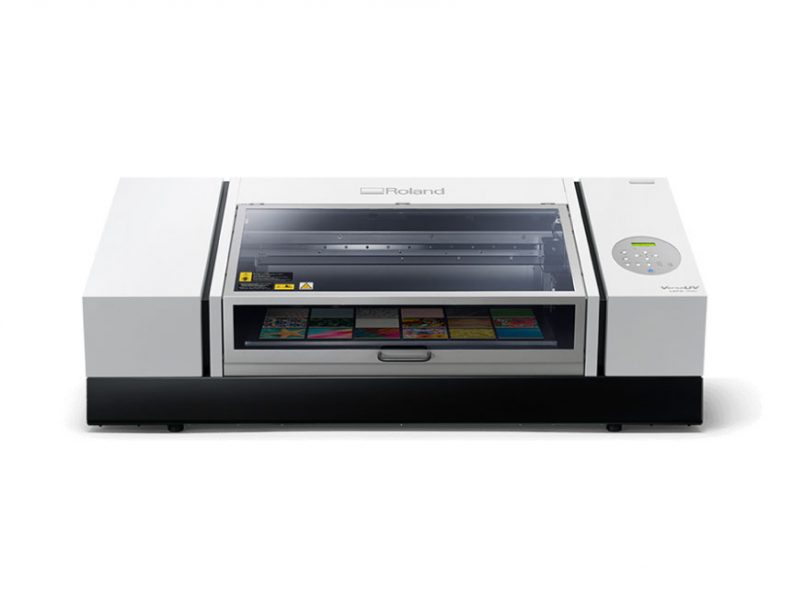 a photograph of a UV-Printer. The printer is a large rectangular grey box. The printer is set against a white backdrop.