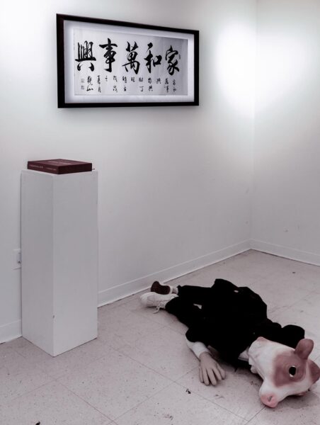 A human figure with a cow's head lies on the floor of a gallery. A framed paper featuring Chinese calligraphy hangs on the wall and a large book is on a pedestal.